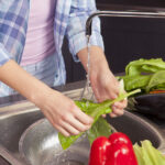 Woman in a blue shirt is washing vegetables in the kitchen sink. Horizontal image. Close-up on hands